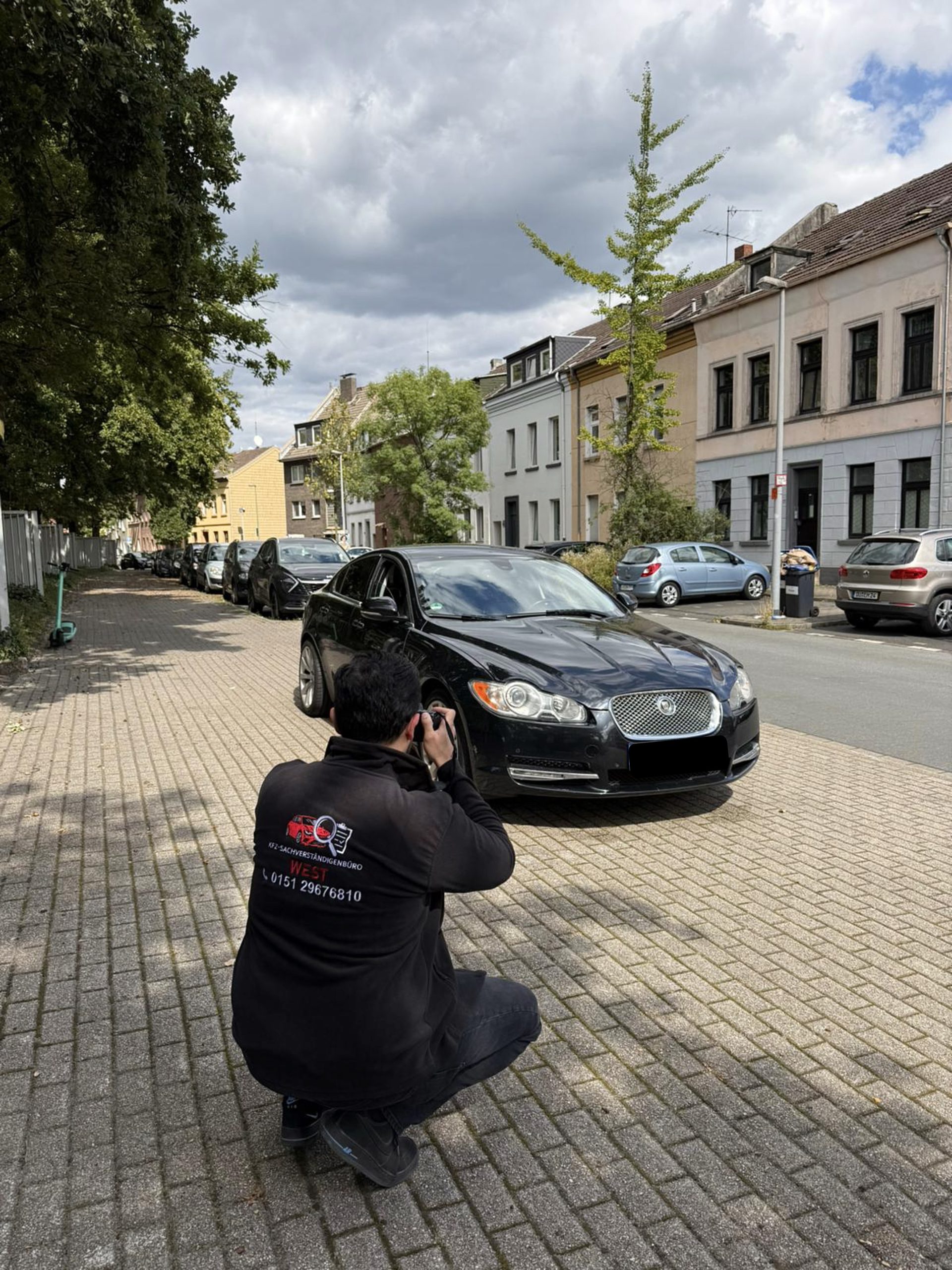 Mann fotografiert schwarzen Jaguar auf ruhiger Wohnstraße mit parkenden Autos und Häuserzeile
