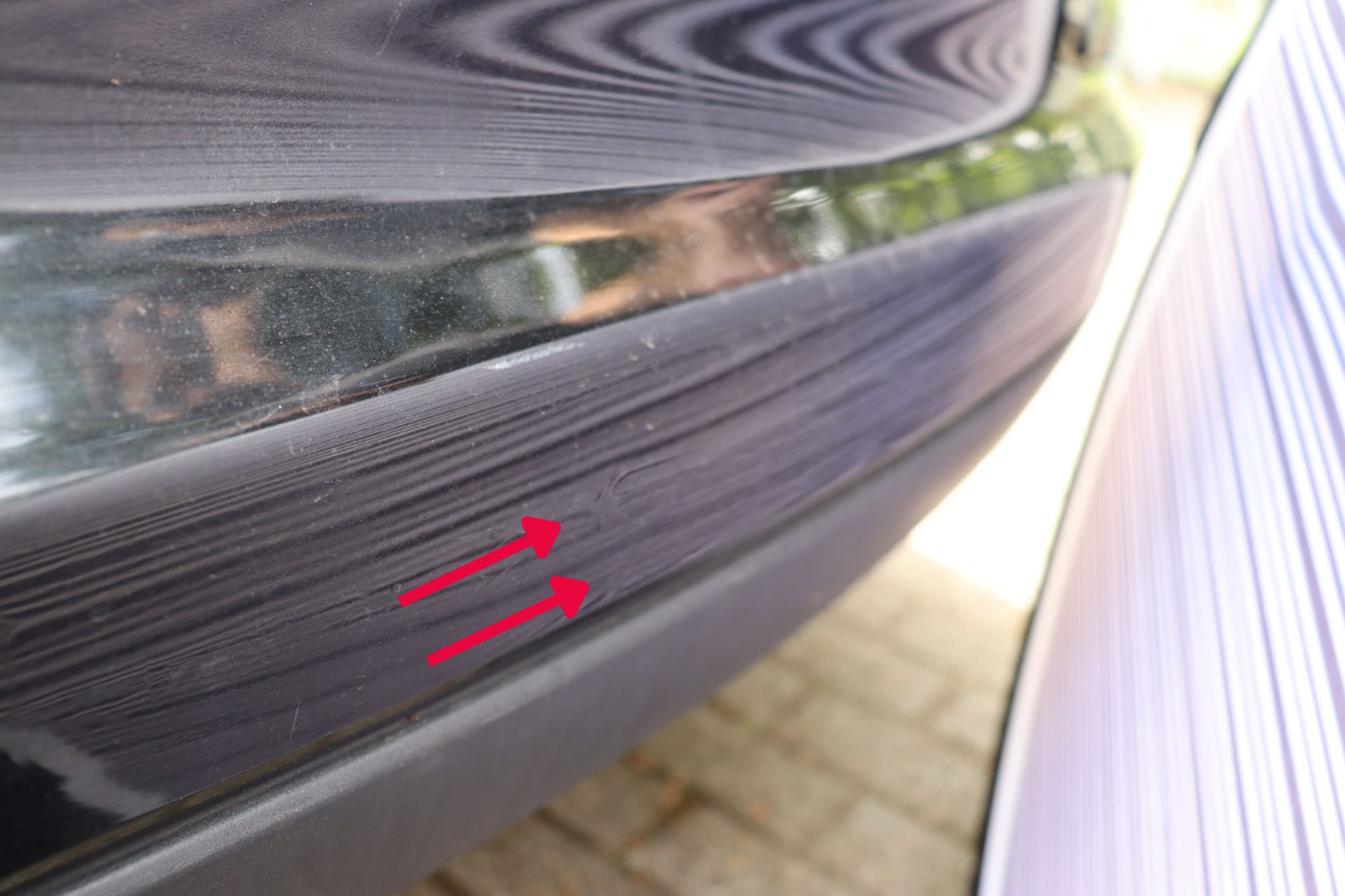 Nahaufnahme von Kratzern und Lackschäden am schwarzen Autostoßfänger, markiert mit roten Pfeilen